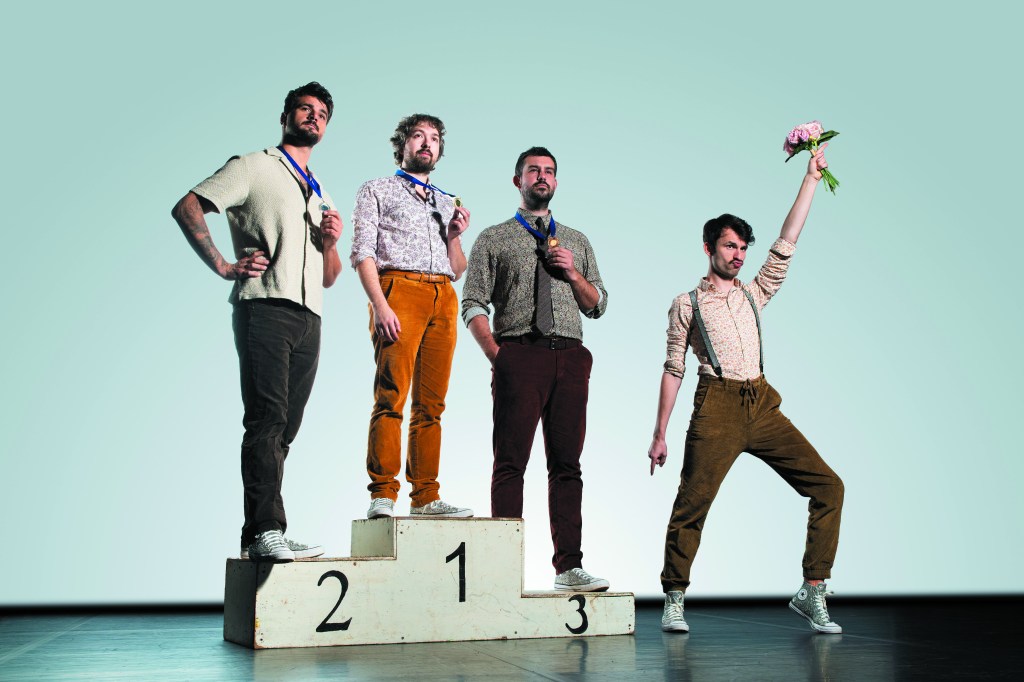 Portrait en noir et blanc du groupe « Les Agités du Bocal ». Les quatre membres posent de manière décontractée et complice devant un mur en briques sombres. Au premier plan, un homme barbu aux cheveux clairs sourit largement. Derrière lui, ses trois acolytes affichent des expressions variées, mêlant sourires et regards malicieux. L'éclairage contrasté met en avant les textures des vêtements (vestes, chemises) et crée une atmosphère à la fois rock et chaleureuse.