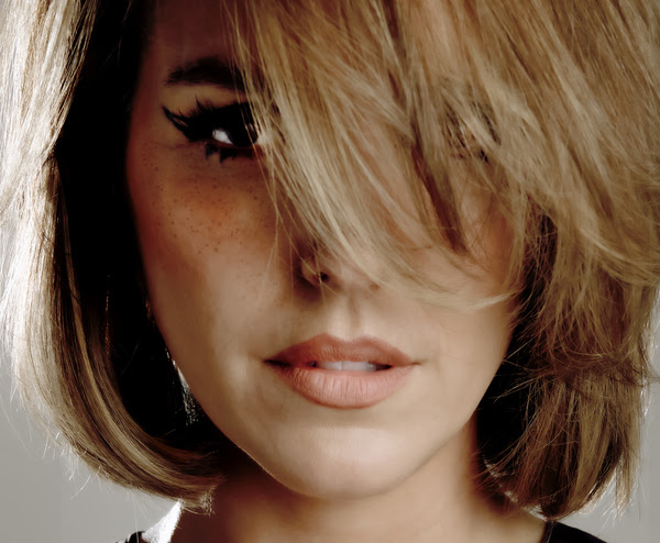 Margaux Simone Un portrait en gros plan de Margaux Simone. Elle a une coupe au carré blond vénitien, avec une mèche de cheveux tombant devant son visage et cachant partiellement son œil gauche. Son regard est dirigé vers l'objectif, souligné par un trait d'eye-liner noir marqué et des taches de rousseur sur ses pommettes. L'éclairage est doux et contrasté, créant une atmosphère à la fois intime et cinématographique.