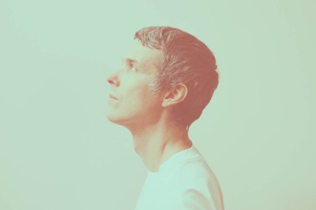 Un portrait de profil de l'artiste Yan Wagner sur un fond uni. Il regarde vers le haut avec une expression pensive. L'image utilise un traitement de couleur bicolore onirique, mêlant des teintes vert menthe pour le fond et des nuances rose saumon pour le sujet, créant une atmosphère douce et éthérée. Il porte un vêtement blanc simple.