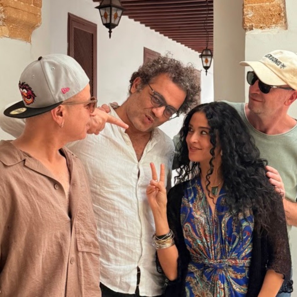 Un portrait de groupe en plan moyen capturant quatre personnes, dont le pianiste Arshid Azarine, sous une arche voûtée blanche de style mauresque. L'ambiance est conviviale et informelle. De gauche à droite : un homme portant une casquette grise et une chemise en lin marron regarde vers le centre. Ensuite, Arshid Azarine, portant des lunettes et une chemise blanche, pose avec un sourire complice. À sa droite, une femme aux longs cheveux bouclés, vêtue d'une robe bleue à motifs paisley et portant de nombreux bracelets, fait un signe de la paix ("peace") en regardant vers Arshid. Enfin, à droite, un homme avec une casquette beige et des lunettes de soleil repose son bras sur l'épaule de la femme. Le fond est une cour intérieure avec des murs blancs, des poutres en bois au plafond et des lanternes traditionnelles suspendues. La lumière naturelle est douce.