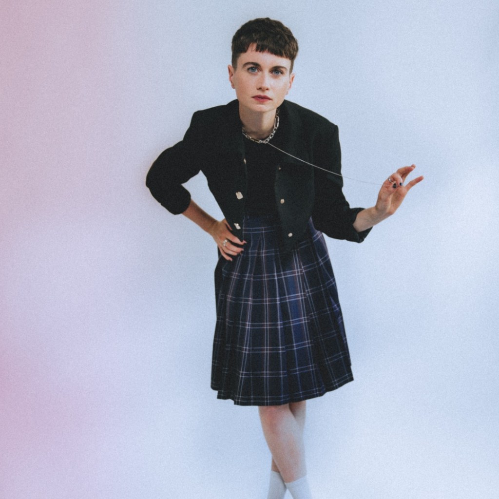 Un portrait en plan moyen de la musicienne Ysé, debout sur un fond de studio blanc avec un subtil voile de lumière rose sur le côté gauche. Elle porte un ensemble structuré et unisexe : un blazer noir ajusté sur un haut assorti, une jupe plissée en tartan bleu marine et vert, et des chaussettes montantes blanches. Sa main gauche repose sur sa hanche, tandis que sa main droite s’élève, tenant une délicate chaîne d’argent fine. Sa coupe courte brune et son maquillage minimaliste mettent en valeur un regard direct et affirmé vers l’objectif. La composition est épurée et contemporaine.